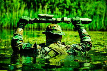 A camouflaged soldier walks through a swamp, submerging himself, his arms and rifle visible, shot...