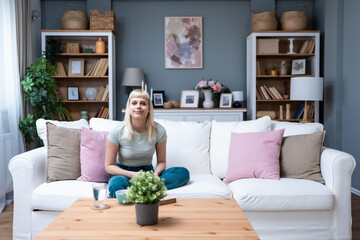 Simple living. Portrait of young beautiful blonde student or college girl sitting at home on sofa...