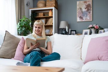 Simple living. Portrait of young beautiful blonde student or college girl sitting at home on sofa...
