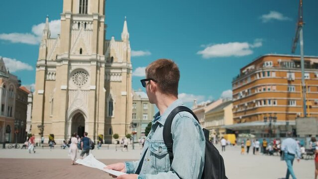 Man tourist with sightseeing map standing outdoors in the old town. Portrait of handsome male traveler backpacker in sunglasses holding map and looking for attractions
