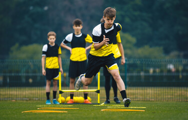Teenage Boy at Training Drill With Young Soccer Coach. Youth Football Player Jumping Over Row of Hurdles at Sports Practice Field. Sports Strength and Agility Training For Footballers
