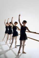 Fototapeta premium Focused preparation. Tender girls, ballet dancers standing at barre and practicing against greys studio background. Concept of ballet, art, dance studio, classical style, youth