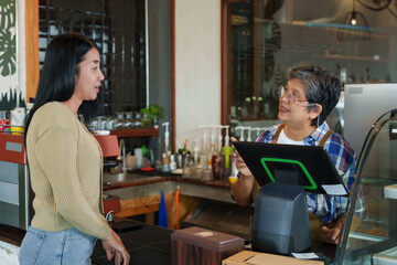 Asian woman converses with barista holding tablet in coffee shop, bread display between them, showing customer service in small business setting.. small family business