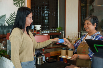 Customer in beige sweater hands over blue credit card to smiling barista in apron, coffee machine and beverages on counter in warm-lit cafe. Cashless payments. small family business