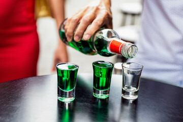 A close-up shot focuses on a hand pouring a shot of strong  liquor into a shot glass, with three...