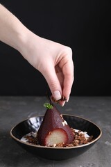 Woman putting muesli on tasty red wine poached pear in bowl at grey table, closeup