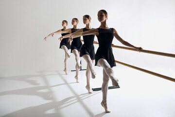 Four elegant girls, ballet dancers standing at barre and warming up, practicing against grey studio background. Focus on discipline. Concept of ballet, art, dance studio, classical style, youth