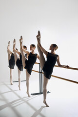 Four ballet dancers line up at barre, performing identical pose with outstretched leg, training against grey studio background. Concept of ballet, art, dance studio, classical style, youth
