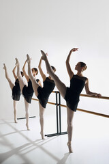 Artistic ballet pose. Four girls, ballet dancers in studio standing at barre, reaching out gracefully with one arm, practicing against grey background. Concept of ballet, art, dance studio, youth