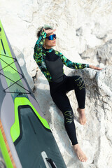 A beautiful sup surfer girl in a wetsuit on the ocean shore against a beautiful blue sky poses sexually with a paddle