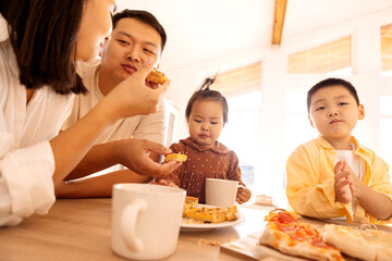 A happy Asian family is eating pizza in the kitchen at home. A young Korean couple and their cute...