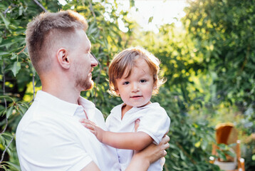 Happy dad holding his cute son in his arms in the garden. Young man and little boy on background of green trees in summer day. - obrazy, fototapety, plakaty