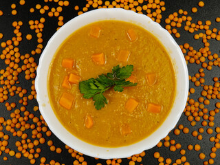 Red lentil puree soup with pumpkin pieces in a white bowl and fresh green parsley are on a black table
 