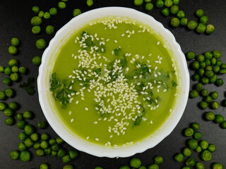 Fresh green pea puree soup with white sesame seeds  in a white bowl and fresh green peas are on a black table