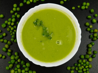 Fresh green pea puree soup in a white bowl and fresh green peas are on a black table.