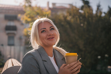 A blonde woman sits on a bench drinking coffee from a yellow cup. She is wearing a gray jacket and...