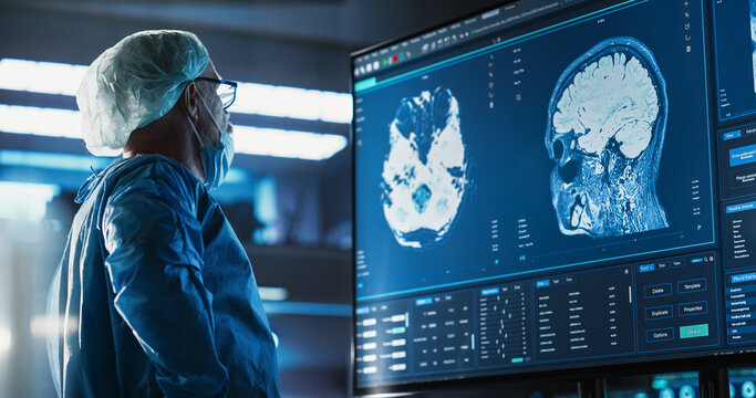 Hospital Research Laboratory: Portrait Of Caucasian Male Neurosurgeon Looking At TV Screen With Brain MRI Scans Of Patient, Analyzing, Preparing For Complicated Neurosurgery, Wearing Surgical Gown.