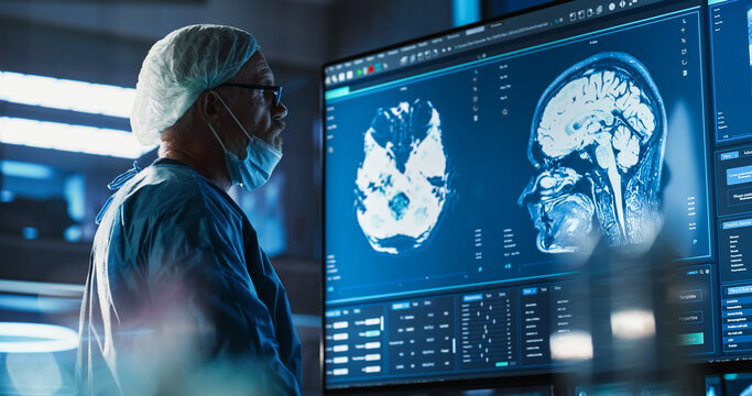 Medical Hospital Research Laboratory: Caucasian Male Neurosurgeon Looking At TV Screen With Brain MRI Scans Of Patient, Analyzing, Preparing For Complicated Neurosurgery, Wearing Surgical Gown.