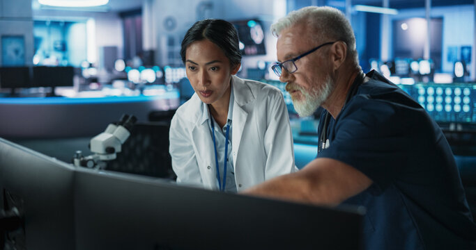 Asian Female Scientist Using Desktop Computer And Talking To Caucasian Male Doctor In Modern Hospital Research Lab. Colleagues Discussing Test Results Of Patients, Treatment Methods, Medication.