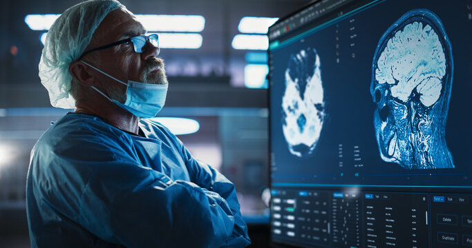Medical Hospital Research Laboratory: Caucasian Male Neurosurgeon Looking At TV Screen With Brain MRI Scans Of Patient, Analyzing, Preparing For Complicated Neurosurgery, Wearing Surgical Gown.