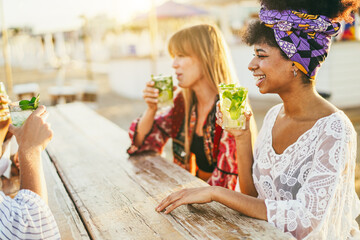 Happy girls drinking mojitos on the beach at chiringuito sunset party - Summer vacation and people lifestyle concept - Main focus on african girl face