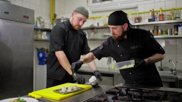 Confident male cook in a black uniform and bandana sprinkles a finished dish with cheese in the kitchen. Duo of chefs in the kitchen preparing a dish together