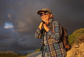Happy senior woman with hat sitting on a hike outdoors in sunset light with dramatic sky ready to...