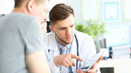 Portrait of concentrated doctor pointing to gadget with finger. Patient listening attentively to...