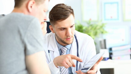 Fototapeta premium Portrait of concentrated doctor pointing to gadget with finger. Patient listening attentively to doc recommendations. Medical treatment concept. Blurred background