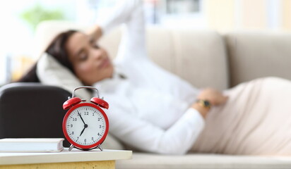 Focus on red alarm clock put near comfortable sofa of tired businesswoman lying on cozy divan and...