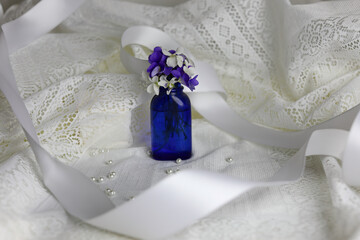 Blue bottle with white and purple violets. White ribbon and lace background.
