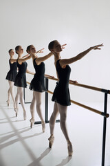 Four ballet dancers, elegant teen girls standing at barre in black costumes and pointe shoes, practicing against grey studio background. Concept of ballet, art, dance studio, classical style, youth