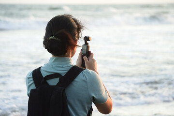 Young asian woman wearing backpack is filming the ocean waves using pocket gimbal camera. Concept...