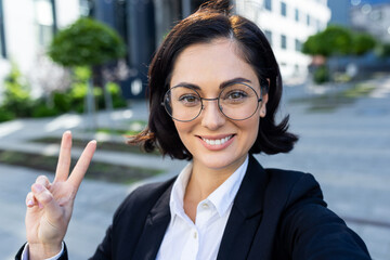 A close-up portrait of a smiling young business woman in a suit and glasses talking on a video call, showing the camera a victory, victory gesture with her fingers