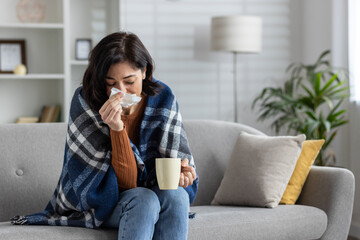 Young woman sitting at home on the sofa and sick, covered with a blanket, wiping her nose from a...