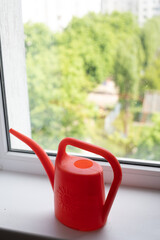 a red watering can on a white windowsill, greenery outside the window