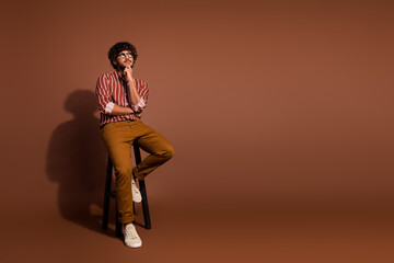 Full size photo of nice young man sit stool look empty space wear striped shirt isolated on brown...