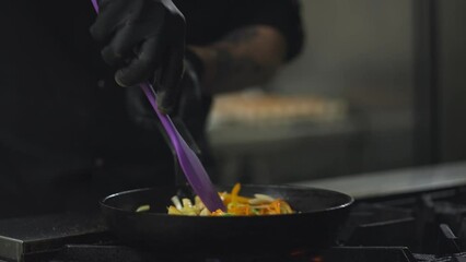 Close-up of a cook in black gloves stirs fried vegetables in a frying pan in a restaurant kitchen. Spectacular cooking in a restaurant kitchen using sparks and fire