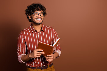 Portrait of nice young man hold book look empty space wear striped shirt isolated on brown color...