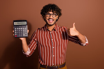 Portrait of nice young man hold calculator show thumb up wear striped shirt isolated on brown color...