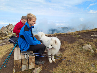Young male hiker in blue jacket smiles at a white dog on a mountain trail