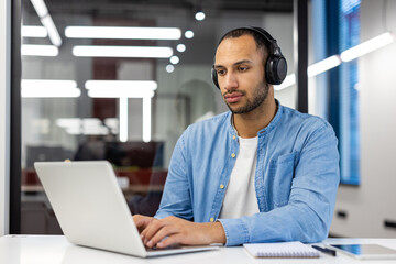 Concentrated young muslim man working on laptop in modern office, sitting at desk wearing headphones