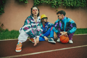 Urban youth fashion. Group of friends sitting in retro style athletic wear outdoor on stadium, with basketball ball. Early 90's outfit trends. Concept of 90s, fashion, youth culture, old-style