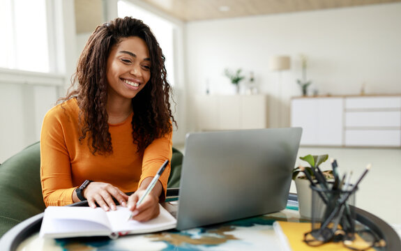Black woman sitting at desk, using pc writing in notebook