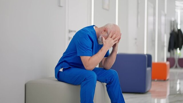 A sad and upset male doctor in a blue uniform an elderly man with glasses and a beard holds his head and cries in the corridor of a modern clinic. An experienced male doctor is upset and worried about