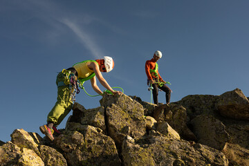 Two people are climbing a mountain, one of them wearing a green harness. The other person is wearing a red jacket - obrazy, fototapety, plakaty