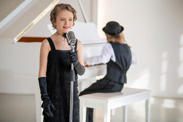 A young girl in a black dress sings into a microphone while a boy plays the piano.