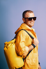 Trendy smiling man in yellow raincoat and sunglasses over blue background.