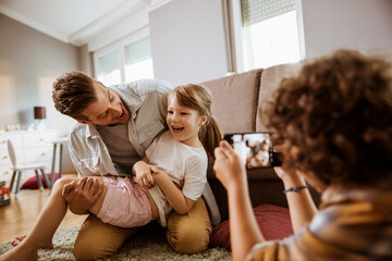Joyful father playing with daughter while son takes photo with smartphone at home