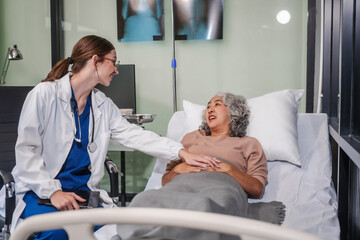 Caucasian female specialist doctor attends to an elderly Asian female patient in a hospital bed,...
